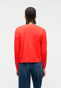 Άτομο με πλεγμένα μαλλιά που στέκεται ανάποδα, φορώντας ένα φωτεινό κόκκινο πουλόβερ και μπλε τζιν τζιν με φόντο ένα απλό φόντο.