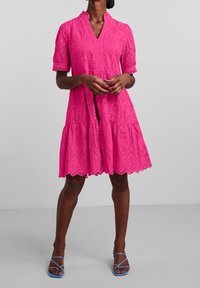 Roze jurk met korte mouwen, een gelaagd ontwerp, oogjespatroon, geschulpte zoom en een V-hals. Gecombineerd met blauwe sandaaltjes met bandjes.