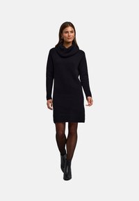 Abito nero a collo alto in maglia con maniche lunghe, caratterizzato da una silhouette aderente e un tessuto testurizzato. Indossato con stivaletti neri alla caviglia.