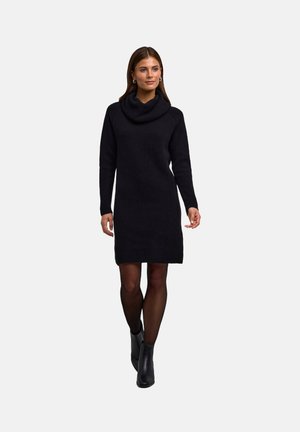Schwarzes gestricktes Kleid mit Kragen und langen Ärmeln, das eine figurbetonte Silhouette und einen strukturierten Stoff aufweist. Getragen mit schwarzen Knöchelstiefeln.