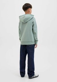 Felpa verde chiaro con cappuccio regolabile, abbinata a jeans larghi blu scuro e sneaker bianco e nero. Tessuto morbido, vestibilità casual.