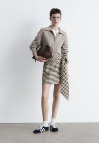 Hellbrauner Strickcardigan, weißes T-Shirt, karierter Rock mit einer gebundenen Detail, dunkelbraune Handtasche und marineblaue Turnschuhe mit weißen Streifen.