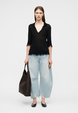 Femme portant un haut noir à col en V, jean large délavé et déchiré, chaussures plates noires, tenant un grand sac cabas tissé sombre, debout devant un fond uni.