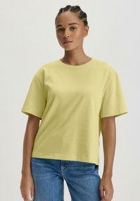 Korte mouwen, losvallend geel katoenen t-shirt met een ronde halslijn. Heeft een gladde textuur en naden bij de mouwen en de zoom.