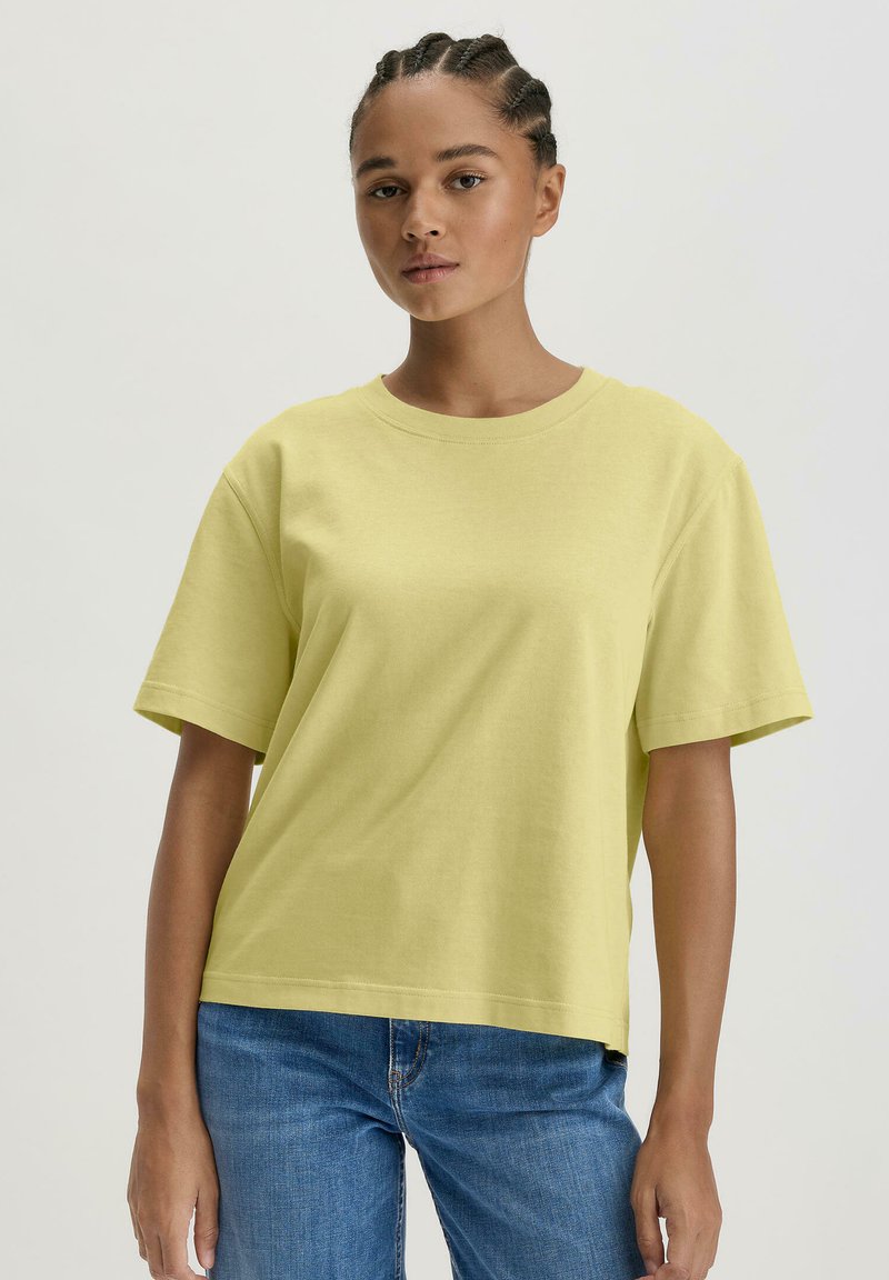 Korte mouwen, losvallend geel katoenen t-shirt met een ronde halslijn. Heeft een gladde textuur en naden bij de mouwen en de zoom.