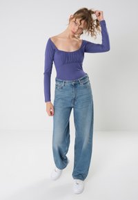 Lilla langermet body med samlet halslinning, parret med høytydende, løstsittende lyseblå jeans og hvite joggesko.