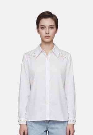 Jeune femme portant une chemise blanche à boutons avec des détails en perles aux épaules et aux poignets, associée à un jean bleu clair, debout sur fond blanc.