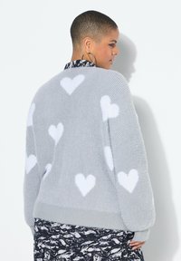 Persona con capelli corti che indossa un maglione grigio peloso con motivi a cuori bianchi e una gonna nera e bianca a fantasia, in posizione laterale.