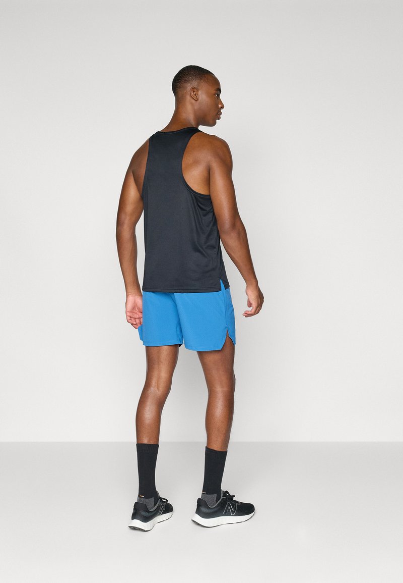 Sportlicher Mann steht, trägt ein schwarzes ärmelloses Oberteil, blaue Shorts, schwarze Socken und schwarze Laufschuhe vor einem schlichten Hintergrund.