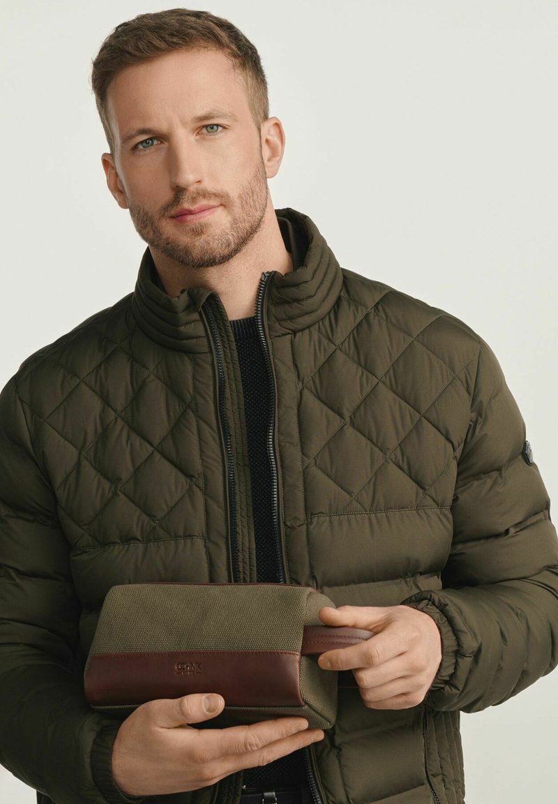 Mężczyzna w pikowanej oliwkowej kurtce trzyma oliwkowo-brązową skórzaną kosmetyczkę na jednolitym jasnym tle.