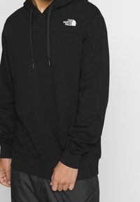 Black hooded sweatshirt made of soft fabric, features a front Kangaroo pocket, drawstrings, and a white logo on the left chest area.