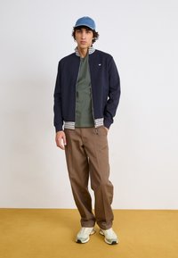 Navy zip-up jacket with checkered collar, olive green t-shirt, brown trousers, and light-colored sneakers against a yellow background.