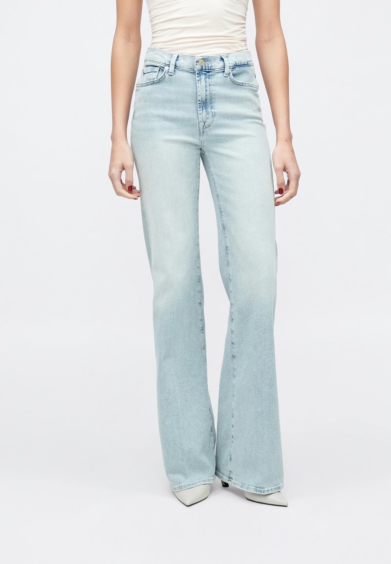 Femme portant un jean large taille haute bleu clair, des chaussures blanches pointues et un top blanc sans manches froncé, debout sur un fond uni.