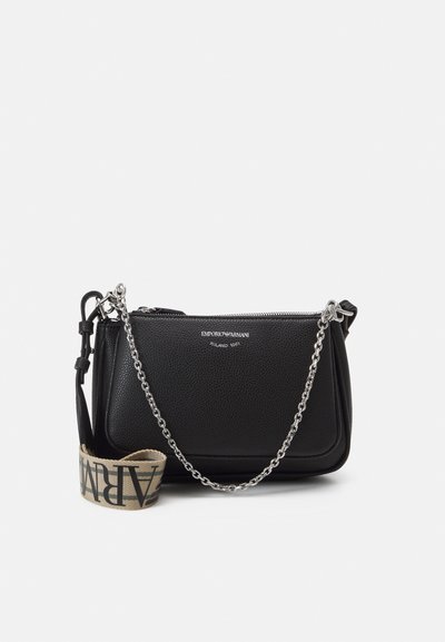 Geantă din piele neagră, tip crossbody, cu o suprafață texturată, curea din lanț argintiu și o curea din material bej pe care este inscripționat numele mărcii.