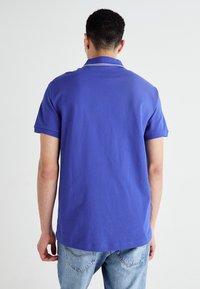 Polo tričko v jednoduchej fialovej bavlne, s krátkymi rukávmi, s kontrastným bielym golierom, v kombinácii s svetlomodrými džínsami.