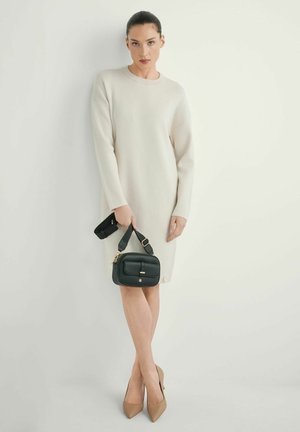 Kobieta w białej sukience z długim rękawem, trzymająca czarną torebkę z szerokim paskiem, ubrana w beżowe szpilki z ostrym noskiem, stojąca na tle białej ściany.