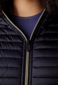 Gros plan d'une veste matelassée noire partiellement zippée sur une chemise bleu marine, montrant une fermeture éclair argentée et des cheveux bouclés au col.