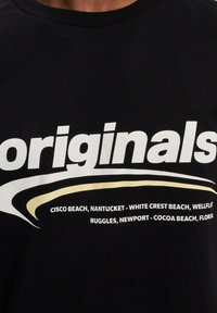 Svart bomull T-skjorte med stor hvit tekst som sier "originals" med en gul bølgekant, og flere strandlokasjoner trykket under.