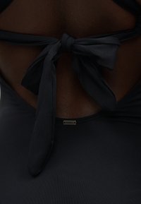 Άτομο με σκουρόχρωμο δέρμα φοράει ένα μαύρο τοπ χιαστί στον λαιμό, δεμένο στην πλάτη με μια μεταλλική ετικέτα της μάρκας O'Neill κάτω από τον κόμπο.