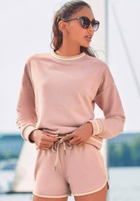 Svietiaco ružová mikina s kontrastným béžovým lemovaním, spárovaná s už zodpovedajúcimi šortkami. Mäkko tkanina, voľný strih a detaily so sťahovacou šnúrkou v páse.