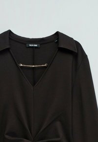 Blusa marrone con scollatura a V e colletto. Presenta un decorazione in catena d'oro sul collo e pieghe sottili sul davanti. Realizzata in un tessuto morbido.