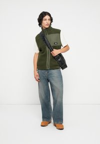 Groene fleece zip-vest met voorzak, witte t-shirt, losse blauwe jeans en bruine instap schoenen, accessorized met een zwarte schoudertas.