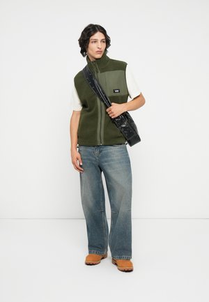Grüne Fleece-Zip-Weste mit Fronttasche, weißes T-Shirt, lockere blaue Jeans und braune Slipper, ergänzt mit einer schwarzen Schultertasche.