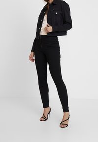 Veste en denim noir, coupe courte, deux poches avant, associée à un jean skinny noir taille haute et des talons noirs à brides.