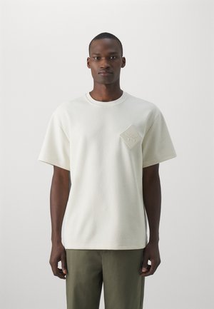Kræmfarvet kortærmet sweatshirt med en tekstureret overflade, der har et diamantformet mærke på brystet med prægede detaljer.