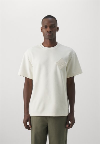 Sudadera corta de manga corta en color crema con un acabado texturizado, que presenta un parche en forma de diamante en el pecho con detalles en relieve.