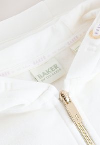Col de veste blanc avec une étiquette « Baker by Ted Baker » et un curseur de fermeture éclair doré embossé de la lettre « B » sur le devant.
