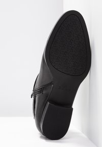 Bottine en cuir noir vue de côté, montrant la semelle texturée avec un motif répété "DKNY" et une fermeture éclair latérale.