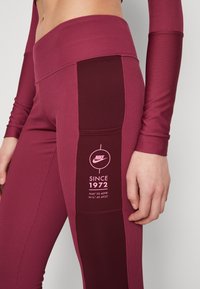 Vinröda leggings i ribbad tyg med en kontrasterande mörk panel och rosa Nike-logotyp. Har en grafik med "SINCE 1972".