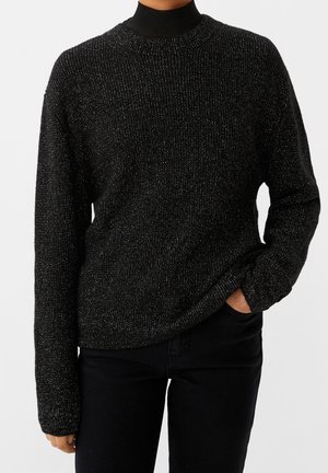 Persoon draagt een zwarte glinsterende gebreide trui over een zwarte coltrui, met één hand in de zak van een zwarte broek.
