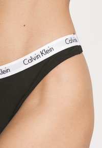 Tanga in cotone nero con un elastico bianco in vita sul quale è stampato in nero "Calvin Klein". Texture liscia e design aderente.