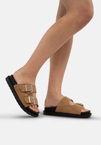 Sandales à glissière en daim marron avec deux sangles réglables en métal argenté, une semelle en caoutchouc noir et un design à bout ouvert.