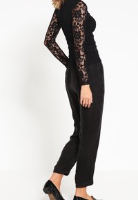 Top in pizzo nero a maniche lunghe con corpetto aderente e maniche trasparenti, abbinato a pantaloni neri larghi e scarpe nere lucide.