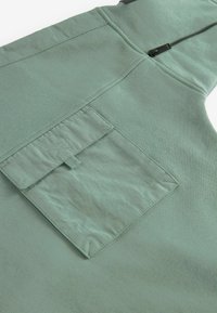 Mintgrön tygväst med en texturerad yta, som har en slät dragkedja fram och en rektangulär ficka på baksidan.