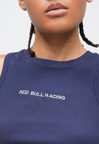 Κοντινό πλάνο ενός ατόμου που φοράει μπλε ριγέ αμάνικο τοπ με το κείμενο «RED BULL RACING» κεντημένο με λευκά γράμματα στο στήθος.