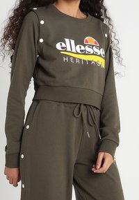 Olivgrön kort sweatshirt med vita tryckknappar på axlarna, med "ellesse HERITAGE" logotyp i vitt och gult. Mjuk textur.