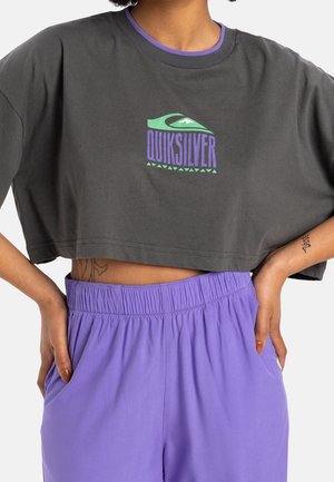 Persoon draagt een cropped donkergrijs Quiksilver T-shirt over een paarse top en hooggetailleerde paarse broek met handen in de zakken.