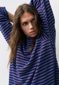 Giovane donna con capelli castani lunghi che indossa un maglione a righe blu e marrone, con le braccia sollevate dietro la testa, che guarda di lato.