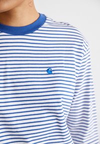 Randig långärmad t-shirt med blåa ränder på vit bakgrund, med en blå cirkulär logotyp broderad på bröstet. Tillverkad av bomull.