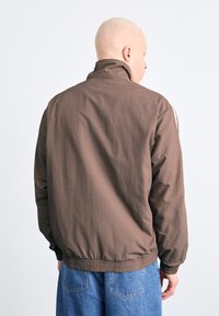 Brun lett jakke med høy krage, elastisk midje og lange ermer. Glatt tekstur med subtil glans, minimalistisk design.