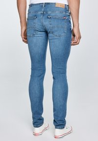 Mees, kes kannab helesiniseid kitsaid pükse ja valgeid tossusid, seisab seljaga vaataja poole tavalise heleda tausta ees.