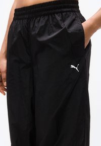 Κλείσιμο εικόνας ενός ατόμου που φοράει μαύρο παντελόνι φόρμας Puma με ελαστική μέση και χέρια στις τσέπες, δείχνοντας το λογότυπο Puma στο μηρό.