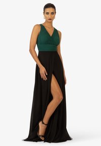 Langes Abendkleid mit einem grünen, plissierten Oberteil und einem fließenden schwarzen Rock mit einem hochgeschlitzten Bein, kombiniert mit schwarzen High Heels.