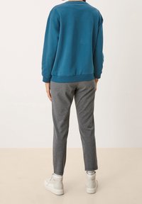 Felpa in cotone verde acqua con polsini a costine, abbinata a pantaloni grigi a gamba affusolata e sneakers bianche, vista da dietro su uno sfondo chiaro.