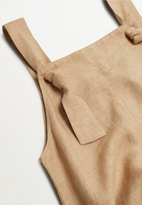Haut sans manches en lin beige avec bretelles nouées, posé à plat sur une surface blanche.