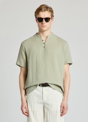 Homme blond portant des lunettes de soleil rondes foncées, une chemise à manches courtes texturée vert clair avec des boutons au col, un pantalon blanc et une ceinture noire.